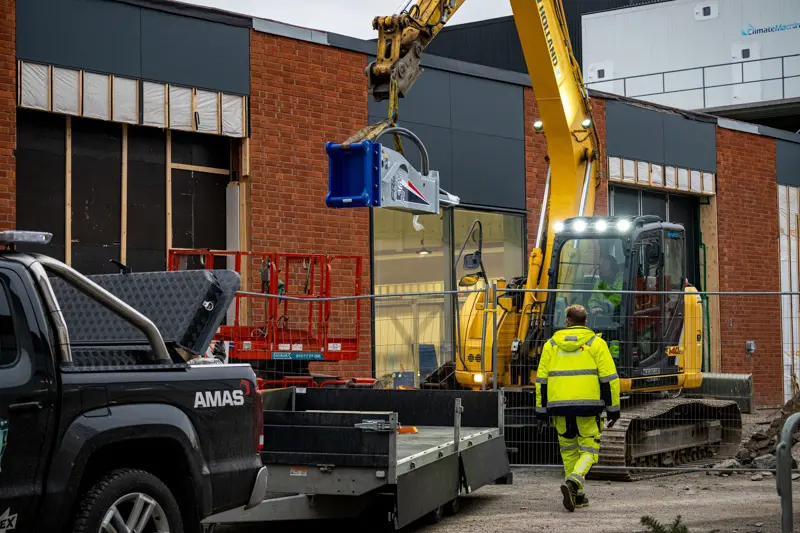 Grävmaskin lastar av en hydraulhammare från släpvagn.