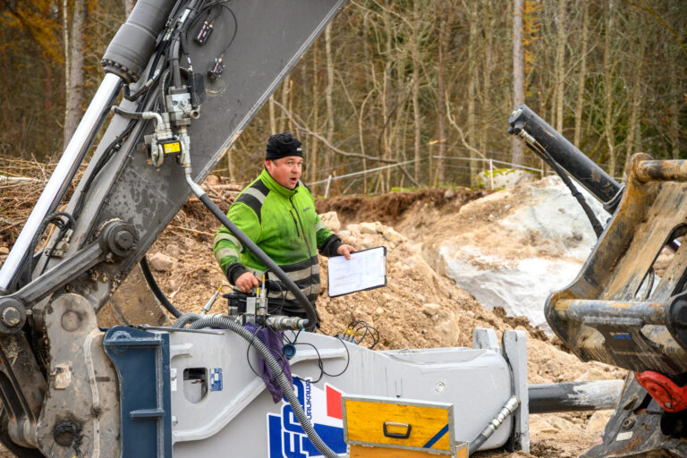 Arbetsledare med manual övervakar hydraulhammarfäste på grävmaskin vid anläggningsarbete utomhus.