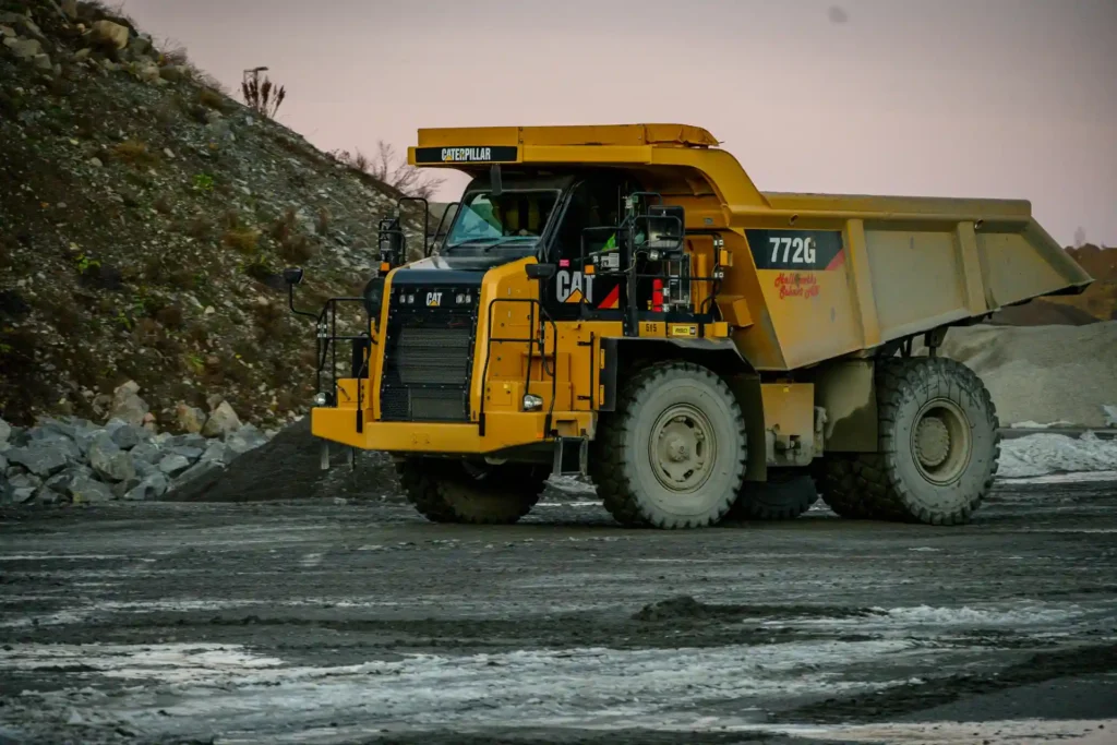 Denna bild visar en stor, gul Caterpillar 772G dumpertruck i profil. Trucken är utformad för att transportera stora mängder material, vilket är vanligt i gruvdrift och byggprojekt. Området runt trucken är en del av en gruvanläggning, med synliga berg och grus på marken. Himlen i bakgrunden har färger som tyder på tidig morgon eller skymning, vilket ger en dramatisk bakgrund till den industriella scenen. Truckens storlek och design antyder att den är avsedd för mycket tunga laster, och det robusta hjulen och chassit är byggda för att hantera tuffa terränger och lastvillkor.