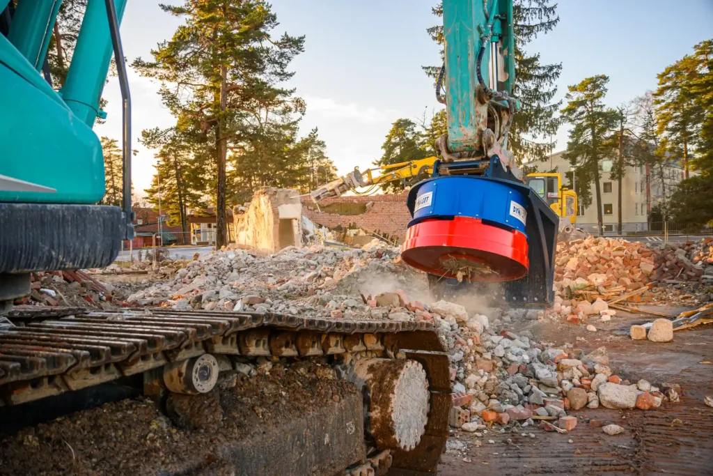 Bilden visar en grävmaskin i arbete på en rivningsplats. Maskinens arm är utrustad med en stor rund skrotmagnet, vilken är i färd med att lyfta eller sortera metallavfall från en hög med bråte som består av bland annat betong och tegelstenar. I bakgrunden syns andra byggmaskiner och en delvis riven byggnad, vilket indikerar att området genomgår en större omvandling eller sanering. Träden och byggnaderna i bakgrunden ger ett sammanhang av platsen, som ser ut att vara i en urban miljö. Dagsljuset och skuggorna antyder att bilden är tagen antingen tidigt på morgonen eller sent på eftermiddagen. Dusten som reses från skrotmagnetens arbete tillför en dynamisk känsla till scenen.