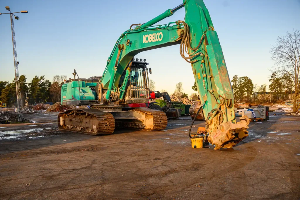 Bilden visar en grävmaskin från märket Kobelco på en byggarbetsplats. Grävmaskinen är av typen bandgrävare, vilket innebär att den har band istället för hjul, vilket gör att den kan manövrera på ojämna ytor och under svårare terrängförhållanden. Maskinen är utrustad med en stor hydraulisk skopa som kan användas för att gräva, lyfta och flytta material. Bakgrunden visar en byggarbetsplats med olika typer av byggmaterial och avfall. Det finns även några träd och en kran i bakgrunden, vilket antyder att platsen är under aktiv konstruktion eller renovering. Liksom den tidigare bilden verkar denna vara tagen sent på dagen, med tanke på ljuset och skuggorna.