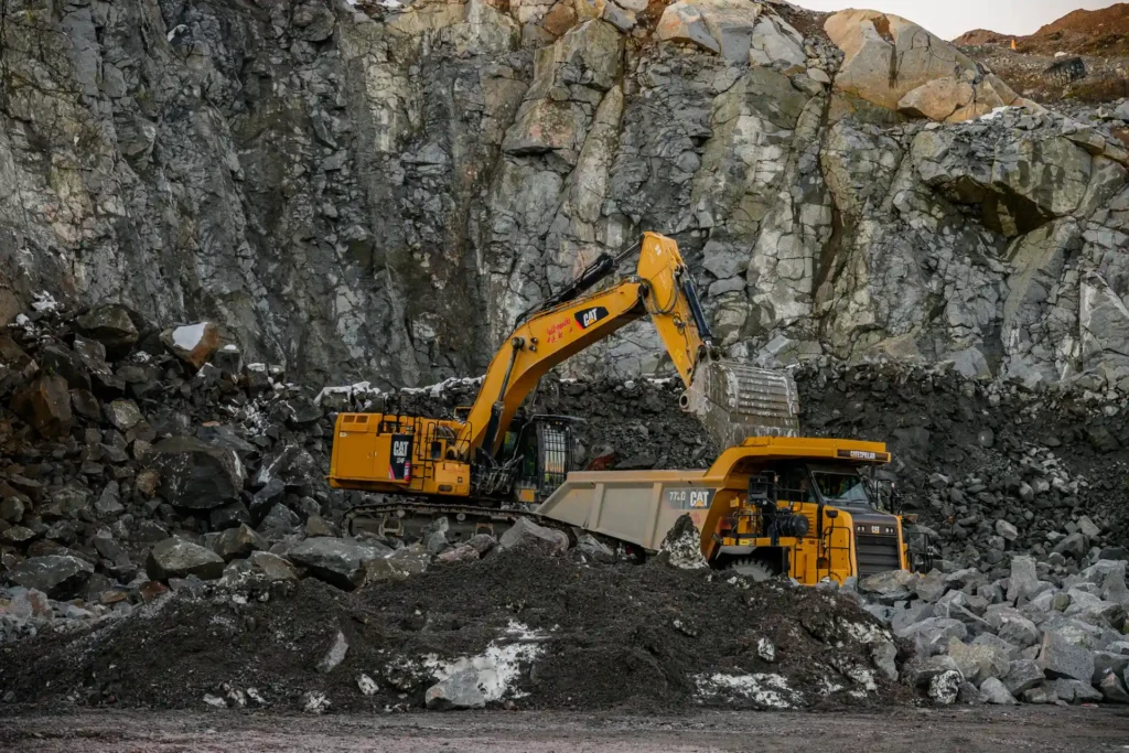 På denna bild ser vi tunga maskiner i arbete på i en bergtäkt. En grävmaskin av märket Caterpillar lyfter just en skopa full med sten och häller innehållet i en stor, gul lastbil, även den från Caterpillar. Detta är en typisk scen från en gruvdrift eller ett stort byggprojekt där material behöver flyttas. Bakgrunden visar en bergvägg med varierande texturer och färger, vilket indikerar sprängning eller grävning som har skett, vanligt i gruvdrift eller stora byggprojekt. Ingen människa syns i denna bild, bara de kraftfulla maskinerna som hanterar det tunga arbetet.
