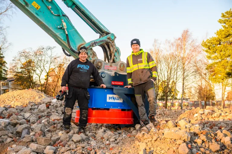Bilden visar två arbetare som står bredvid en del av en grävmaskin med en hydraulmagnet. De bär arbetskläder och skyddsutrustning, vilket är standard för säkerhet på en byggarbetsplats. En av personerna bär på en skruvdragare. De står framför en hög med rivningsavfall, vilket antyder att de kanske arbetar med rivning eller sanering av området. I bakgrunden kan man se träd och en klar himmel, vilket tyder på att fotot är taget utomhus under dagtid. Deras klädsel och utrustning visar att de är professionella inom bygg- eller demolering.