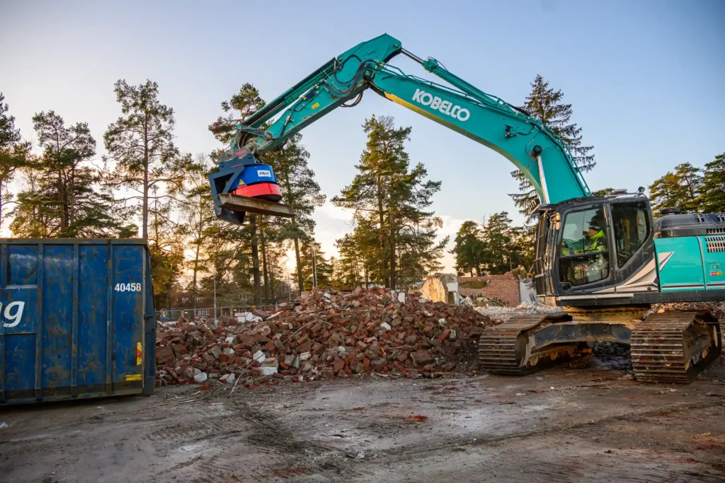Bilden visar en byggarbetsplats där en grävmaskin utrustad med en skrotmagnet lyfter en färgglad metallbalk. Skrotmagneten är fästad vid grävmaskinens arm och använder magnetism för att hålla fast och transportera metallföremål. I bakgrunden syns en hög med rivningsavfall, främst tegelstenar, och några träd som antyder att platsen kan vara i närheten av en skog eller ett skogsområde. Det ser ut att vara sen eftermiddag eller tidig kväll, eftersom himlen har en mild orange ton, vilket kan indikera att solen håller på att gå ner.