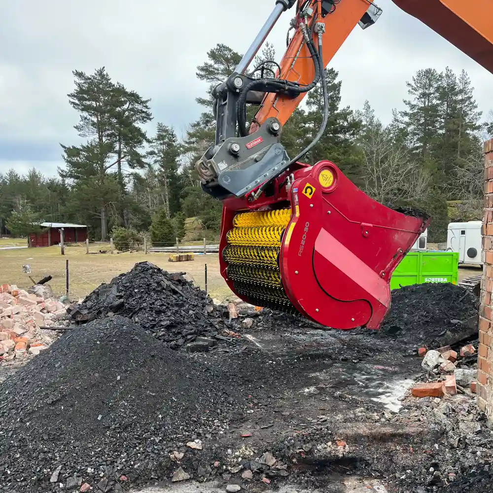 Orange grävmaskin med siktskopa som bearbetar massor efter brand, alltså brandsanering med gröna träd i bakgrunden.