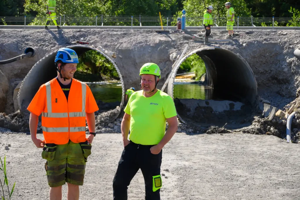 Amos Sjöstedt och Johan Martengård vid Kölstaån under utbyggnaden av E18, med stora trummor i bakgrunden.