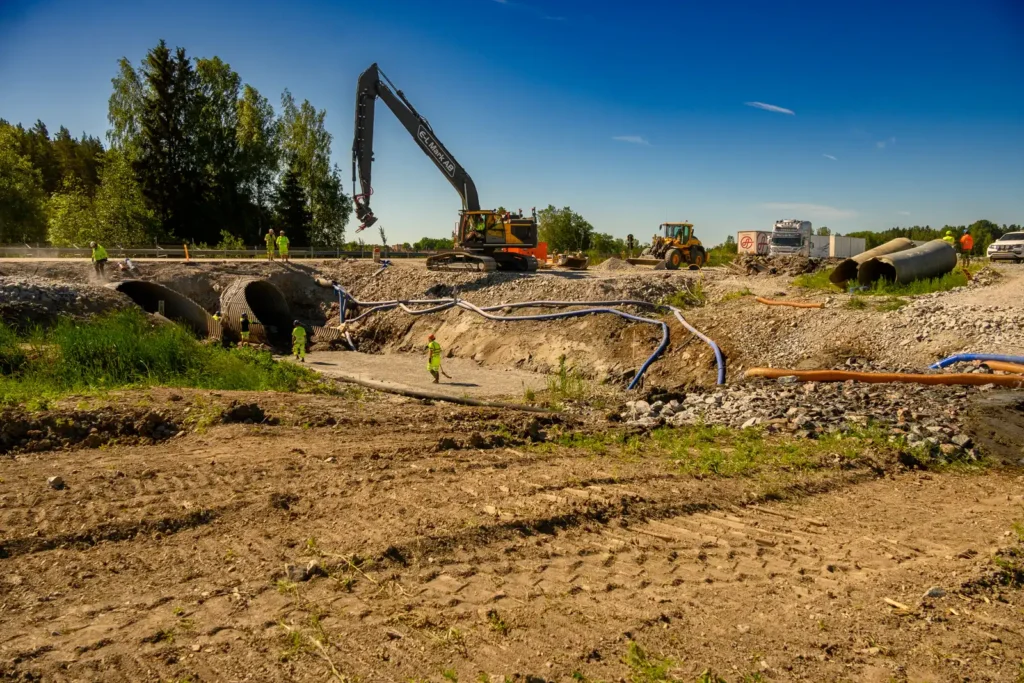 Byggarbete vid Kölstaån under utbyggnaden av E18, med arbetare och maskiner i aktivitet.