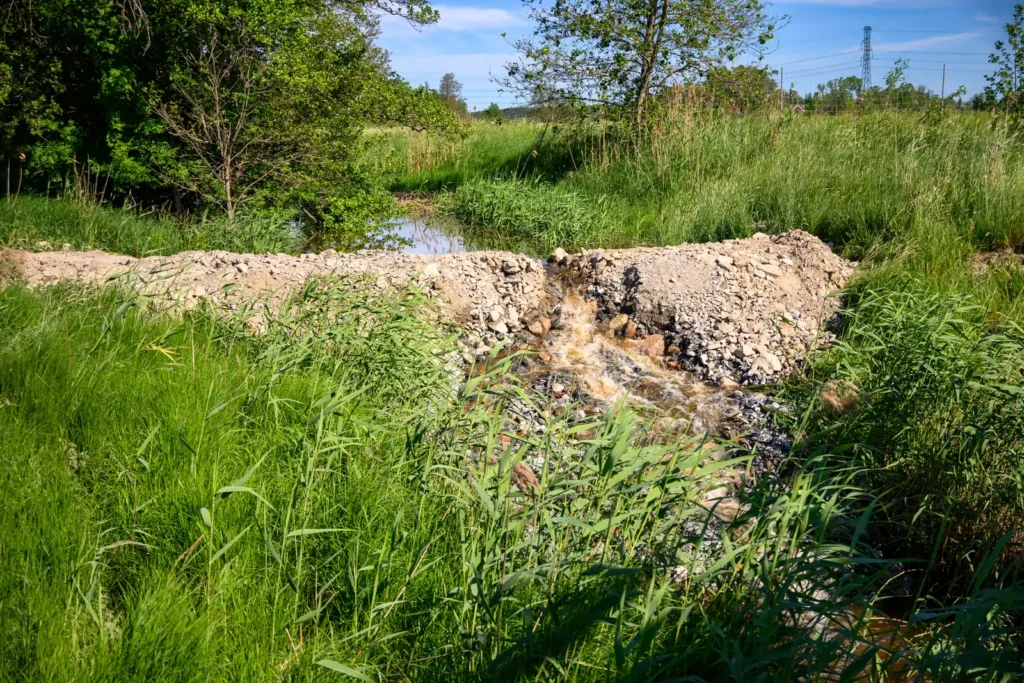 Vattenflöde vid Kölstaån under utbyggnaden av E18, omgiven av grön vegetation och grävningsarbeten.